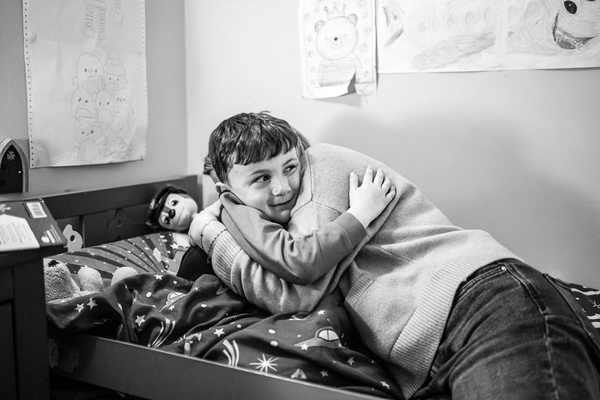 Black and white photo of a smiling child relaxing on a bed with patterned bedding in a cozy bedroom.