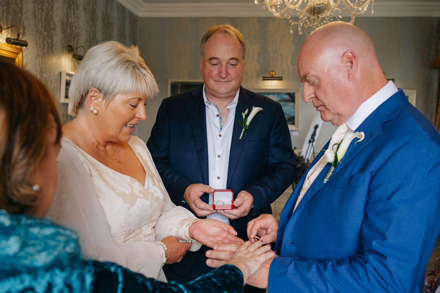 Groom placing wedding ring on brides finger during ring exchange ceremony with guests.
