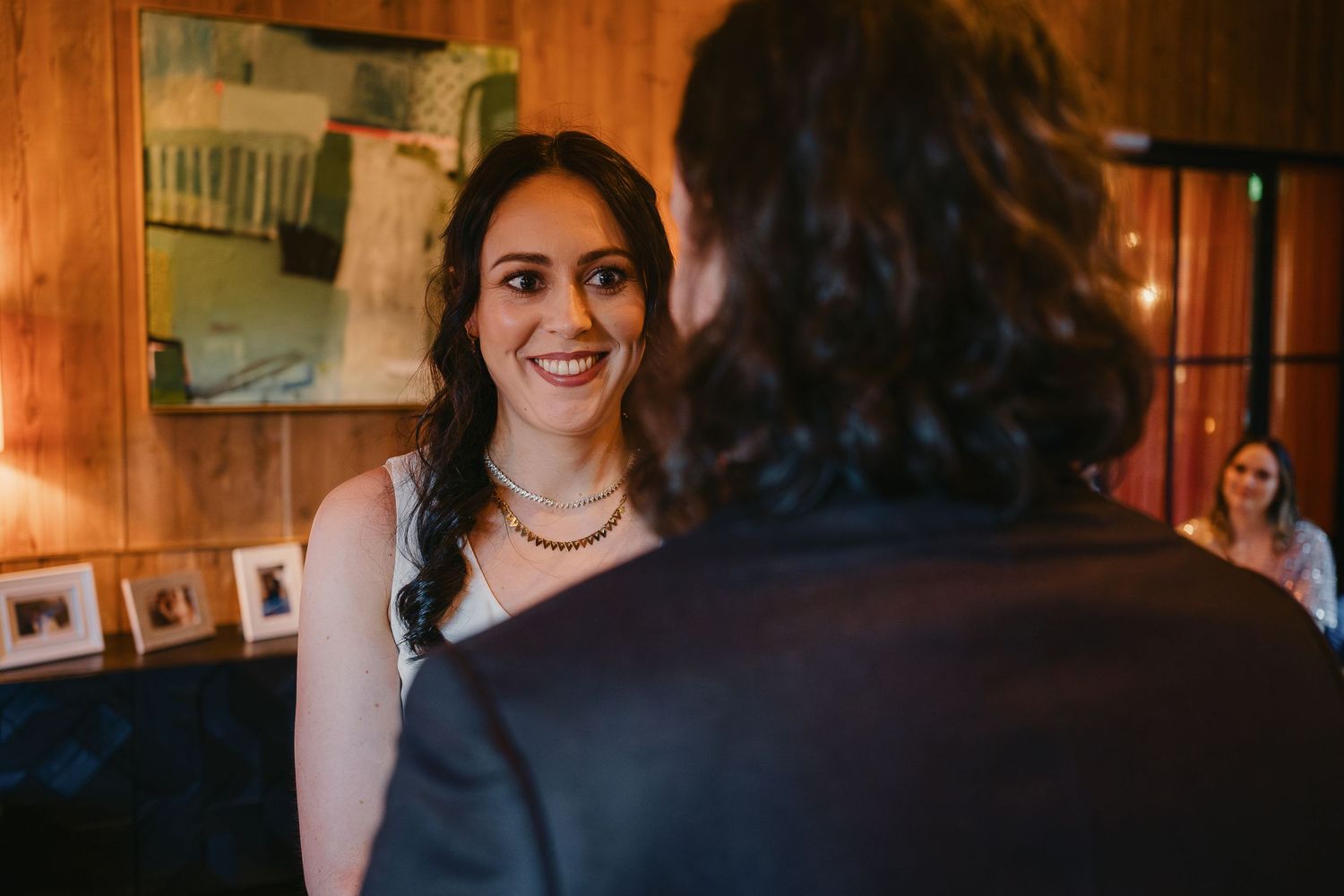 A smiling bride in a white dress wearing a gold necklace talks with a guest inside a warmly lit wooden venue.