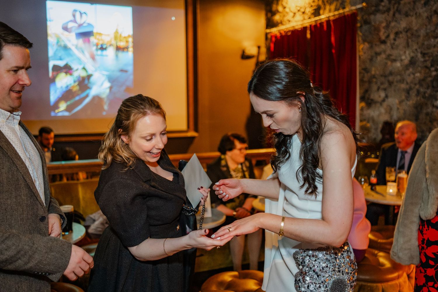 Two women exchanging gifts at an elegant indoor event with guests and decorative lighting in the background.