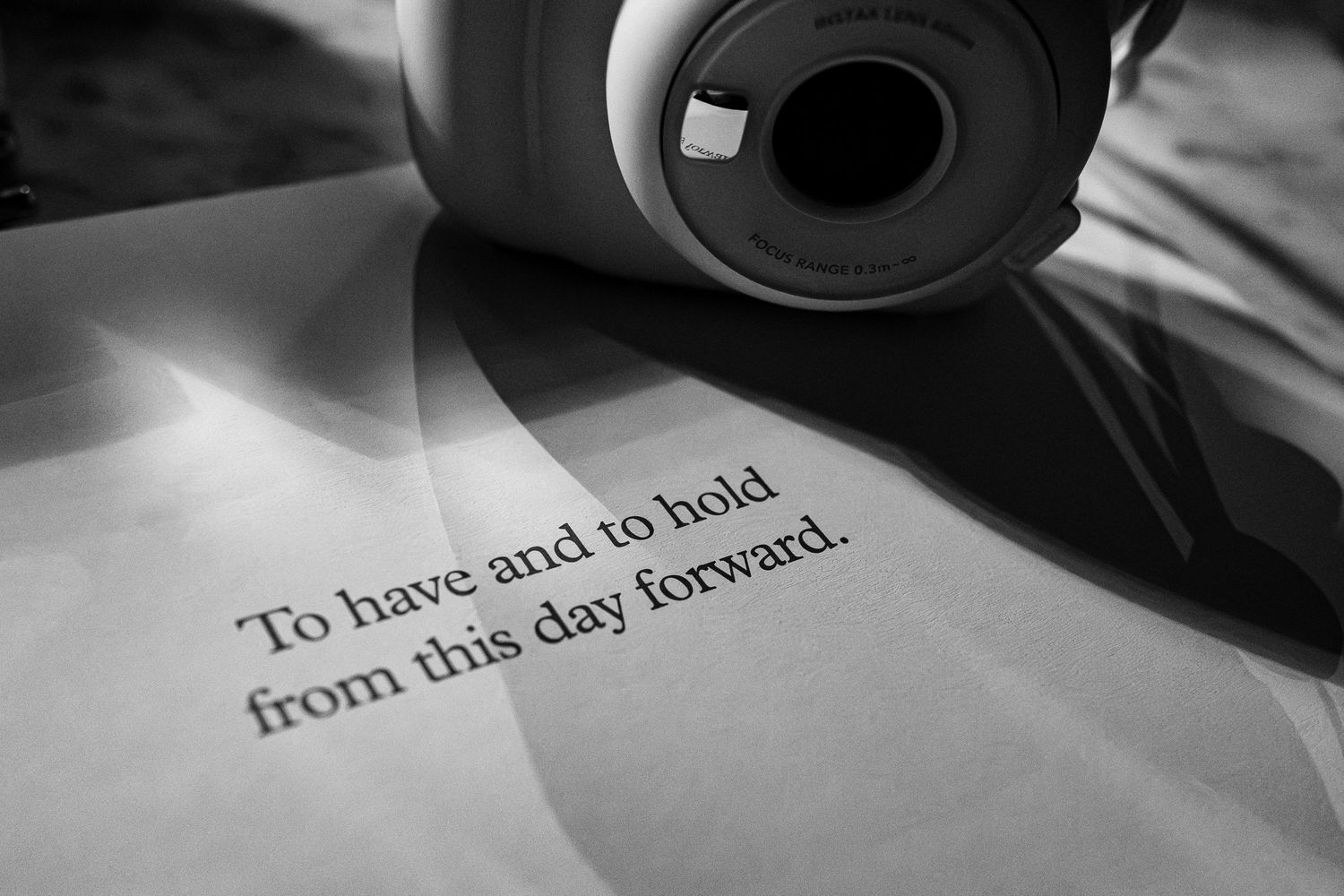 Black and white photo of a Fujifilm Instax camera resting on paper with wedding vow text reading to have and to hold.
