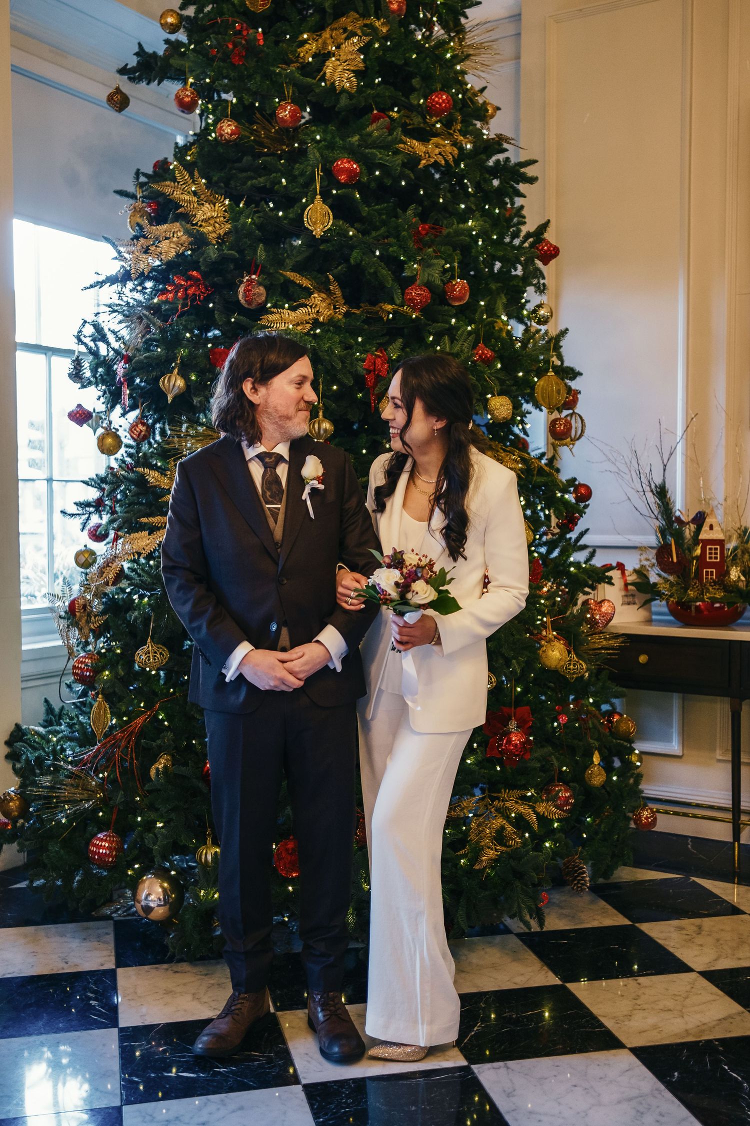 A bride in a white suit and groom in navy hold a bouquet before a decorated Christmas tree on a checkered floor.