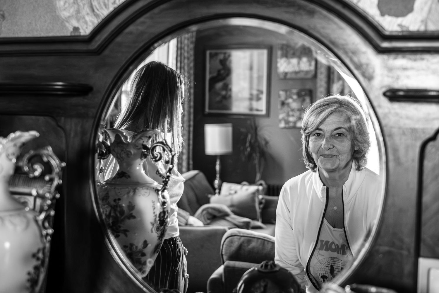 Black and white photo of a woman reflected in an ornate round mirror surrounded by vintage decor.