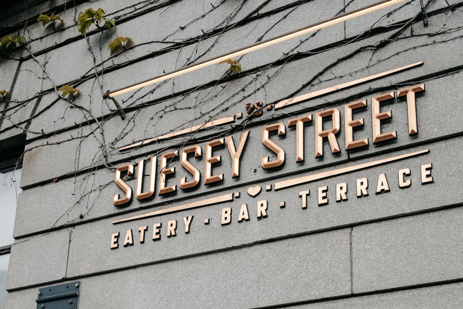 Suesey Street eatery, bar, and terrace sign mounted on a modern stone building facade with bare branches.