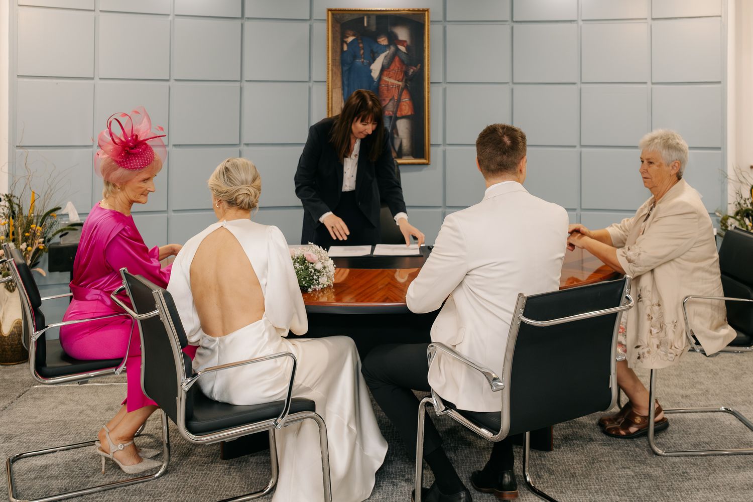 Small wedding ceremony at a conference table with bride, groom, officiant, and two guests in formal attire.