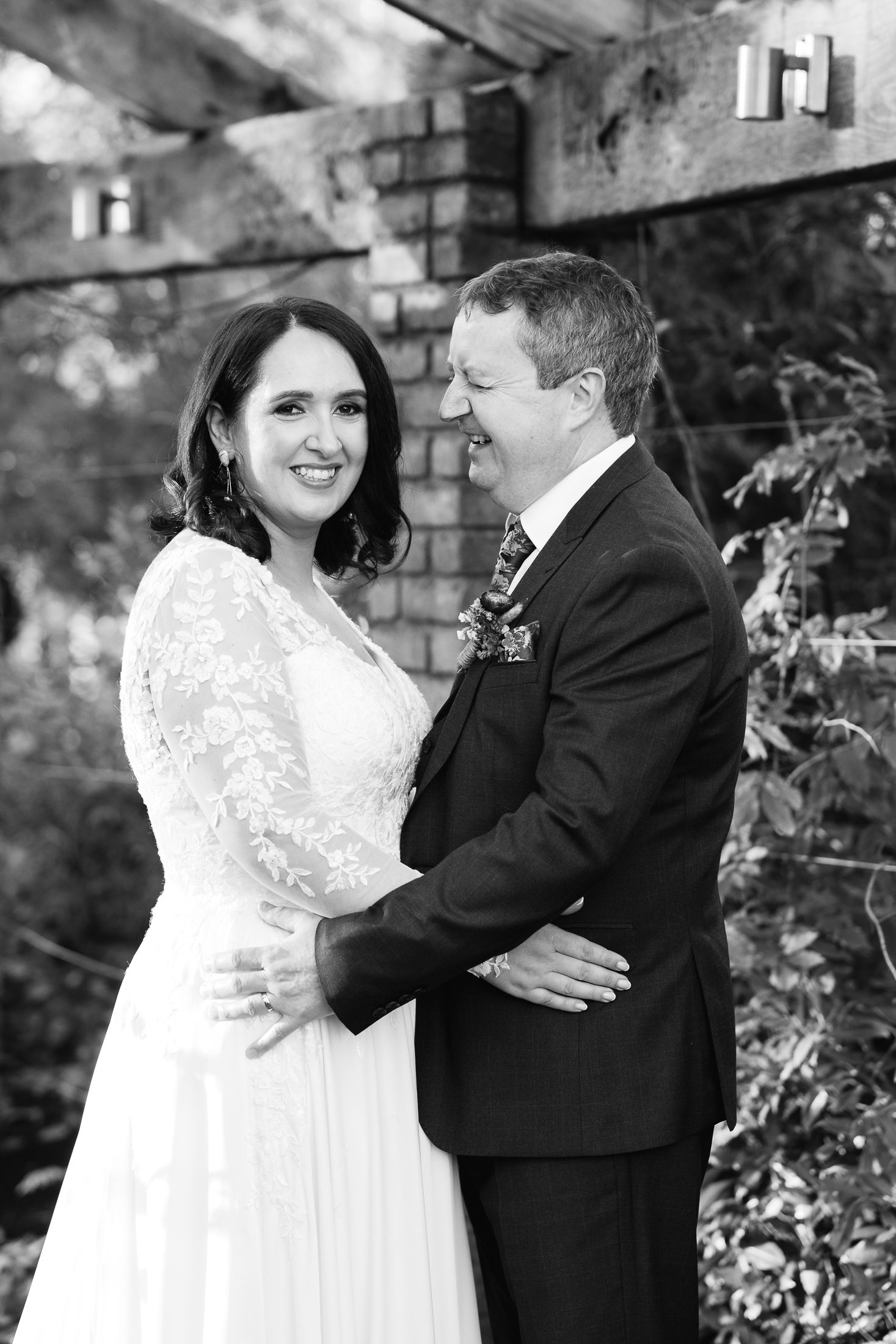 Black and white wedding portrait of smiling bride in lace gown and groom in suit embracing outdoors.