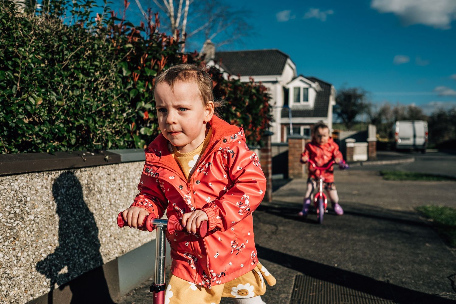 A child in a bright red jacket plays outside on a sunny residential street.
