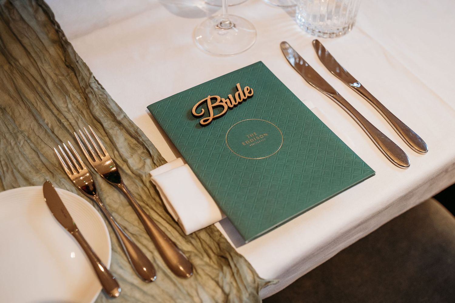 A green menu book labeled 'Bride' on a formal place setting with gold cutlery.