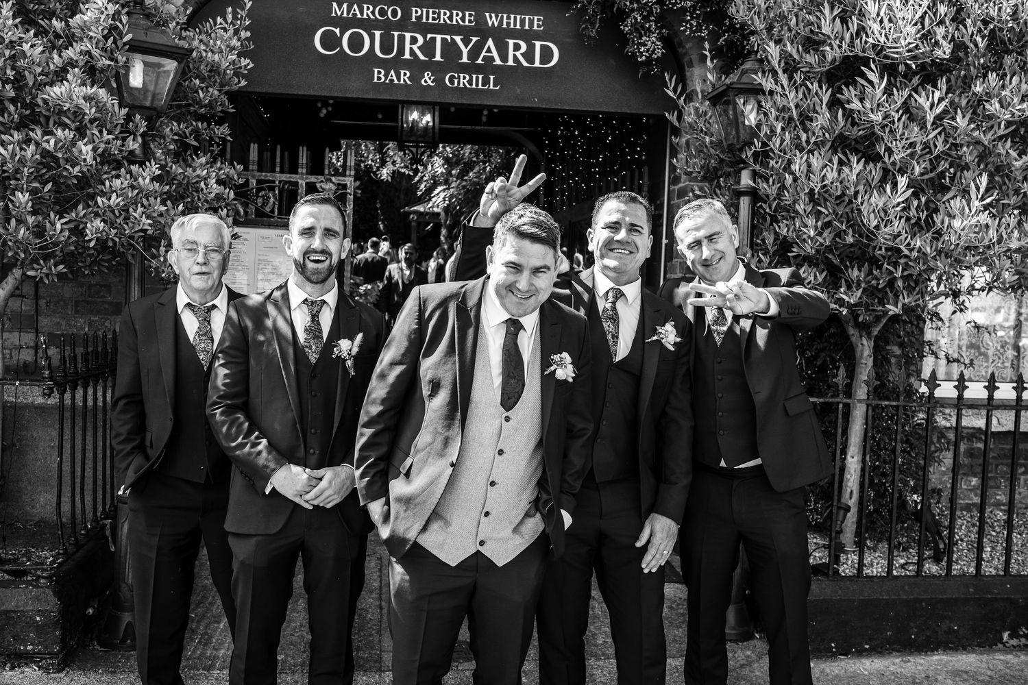 Groomsmen in dark suits pose together outside Marco Pierre White Courtyard.