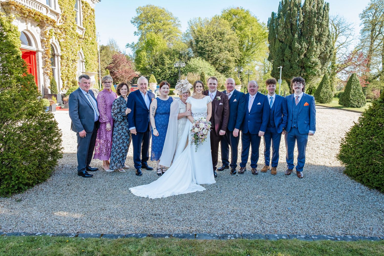Wedding party stands together outside on a gravel path surrounded by green trees and stone buildings.