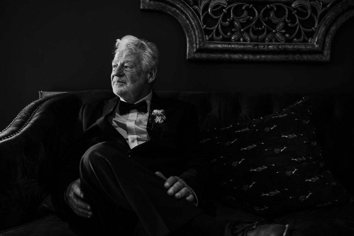 Dramatic black and white portrait of person in tuxedo seated on ornate couch.