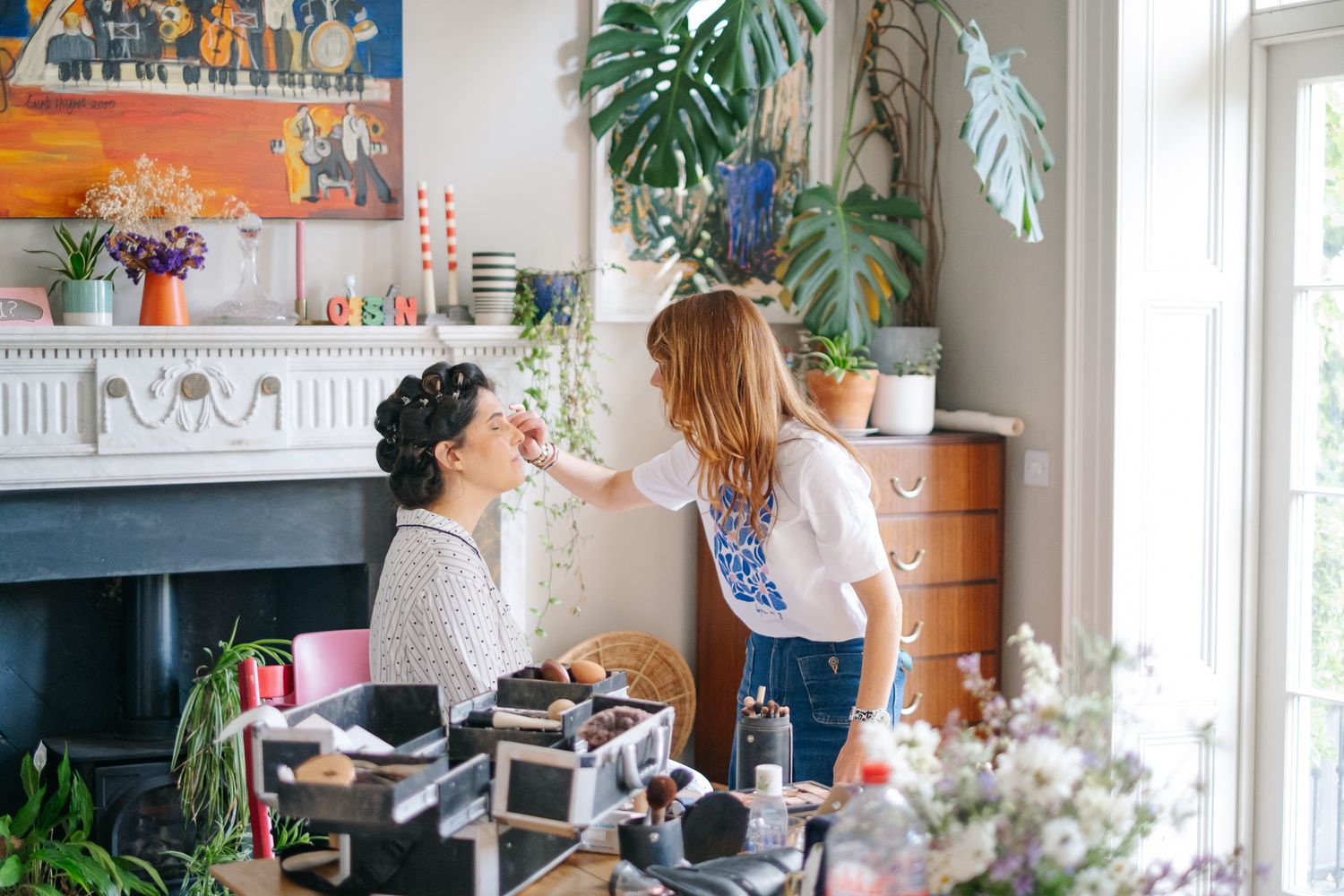 Makeup artist applying makeup to a seated woman in a bright, plant-filled room with a fireplace and colorful artwork.