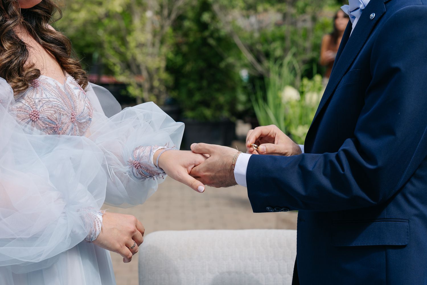 Groom in navy suit placing wedding ring on bride's finger during outdoor ceremony.