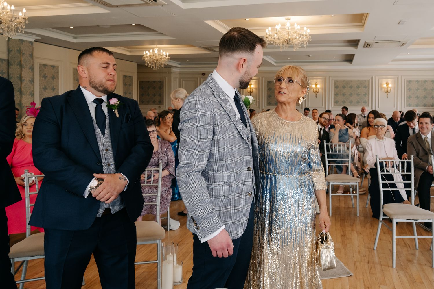 Elegantly dressed wedding guests stand in a grand ceremony hall with chandeliers and white chairs.