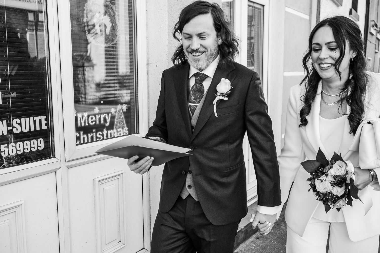 Happy bride and groom laughing outside building with Merry Christmas sign in black and white photo.