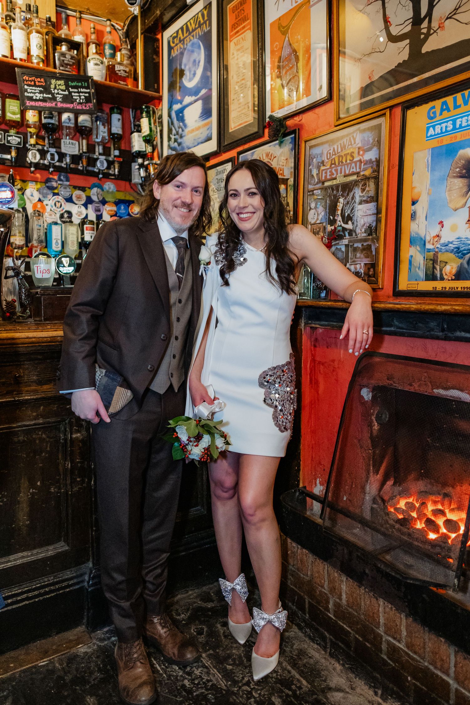 Newlywed couple posing by a cozy pub fireplace, bride in white mini dress holding a bouquet, groom in brown suit.