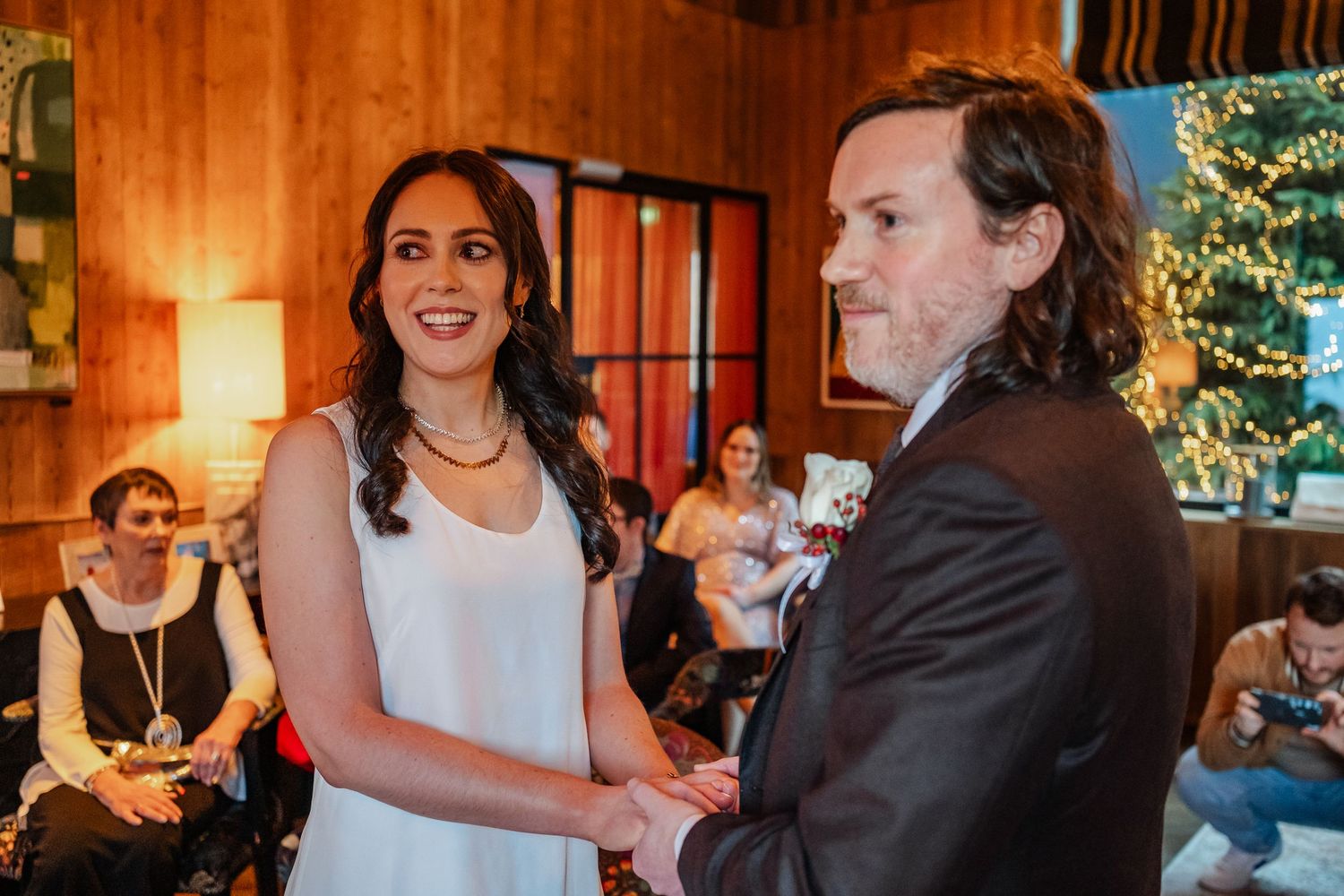 Bride and groom holding hands smiling at indoor wedding ceremony with Christmas tree.