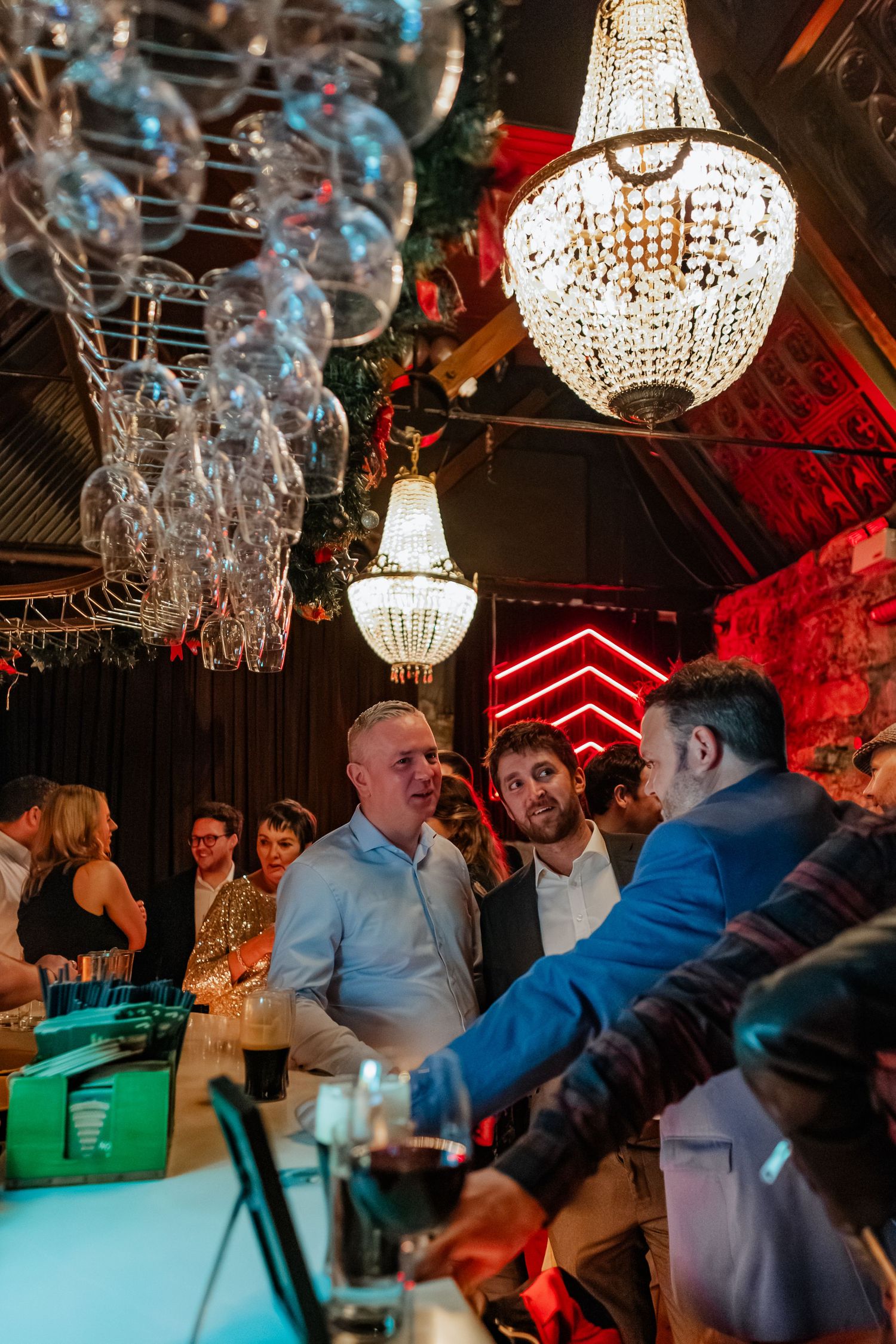 People socializing at a bar with crystal chandeliers and wine glasses overhead in a festive venue.