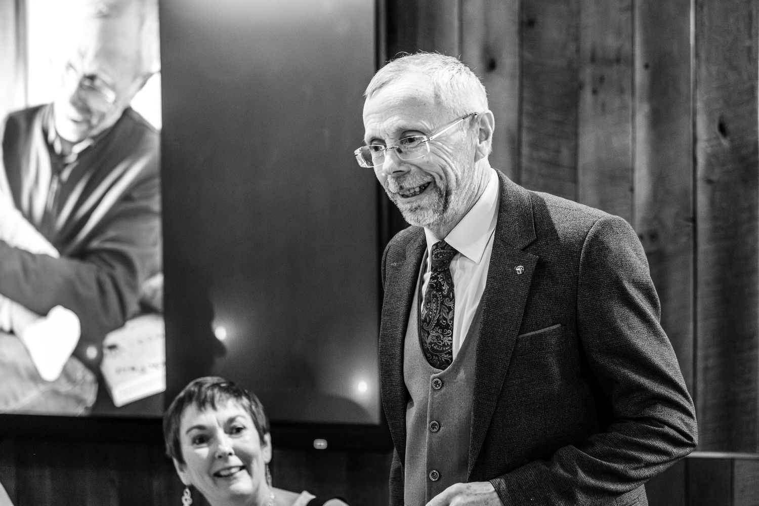 Elderly man smiling warmly at an indoor event, wearing glasses and a suit, with a woman seated behind him.