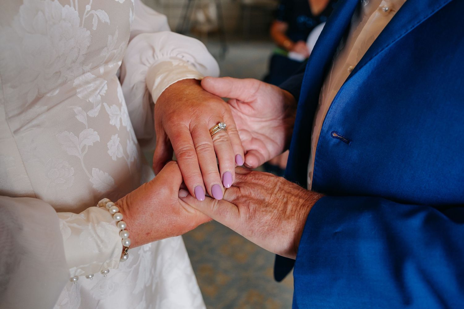 Close-up of hands exchanging wedding rings during a marriage ceremony, with a pearl bracelet visible.
