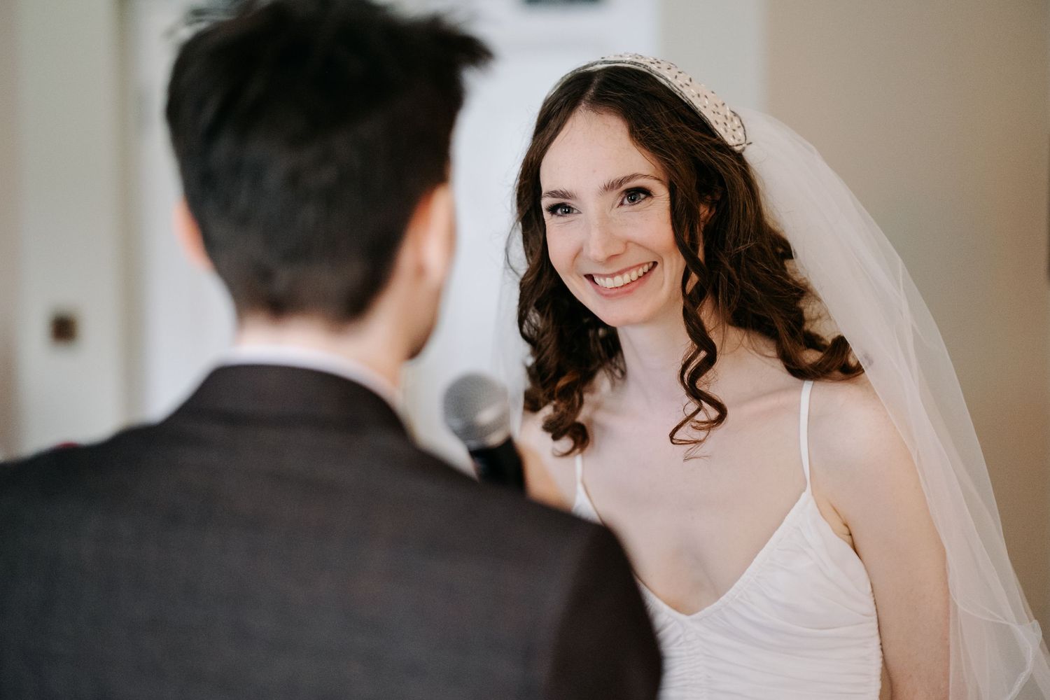 Bride in white gown and veil smiling at groom during wedding ceremony.