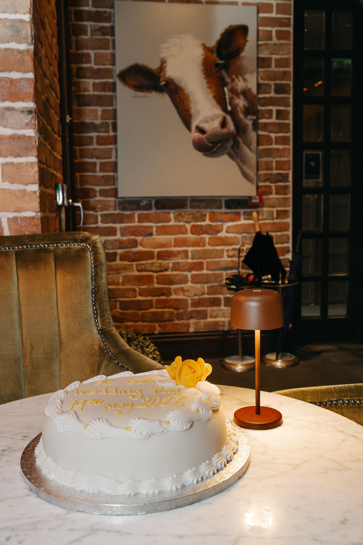 White birthday cake with yellow roses on marble table, cow painting on brick wall in elegant restaurant setting.