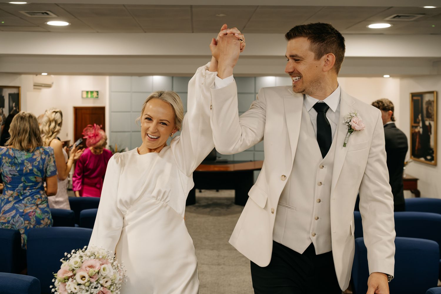 Joyful newlyweds celebrate after wedding ceremony, bride in white gown holding bouquet, groom in white suit raising hands.