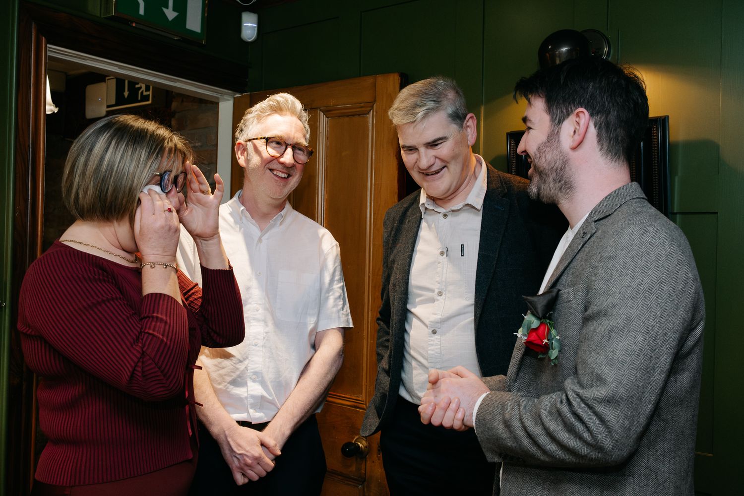 Four people socializing indoors, smiling and shaking hands at a formal gathering in a green-walled venue.