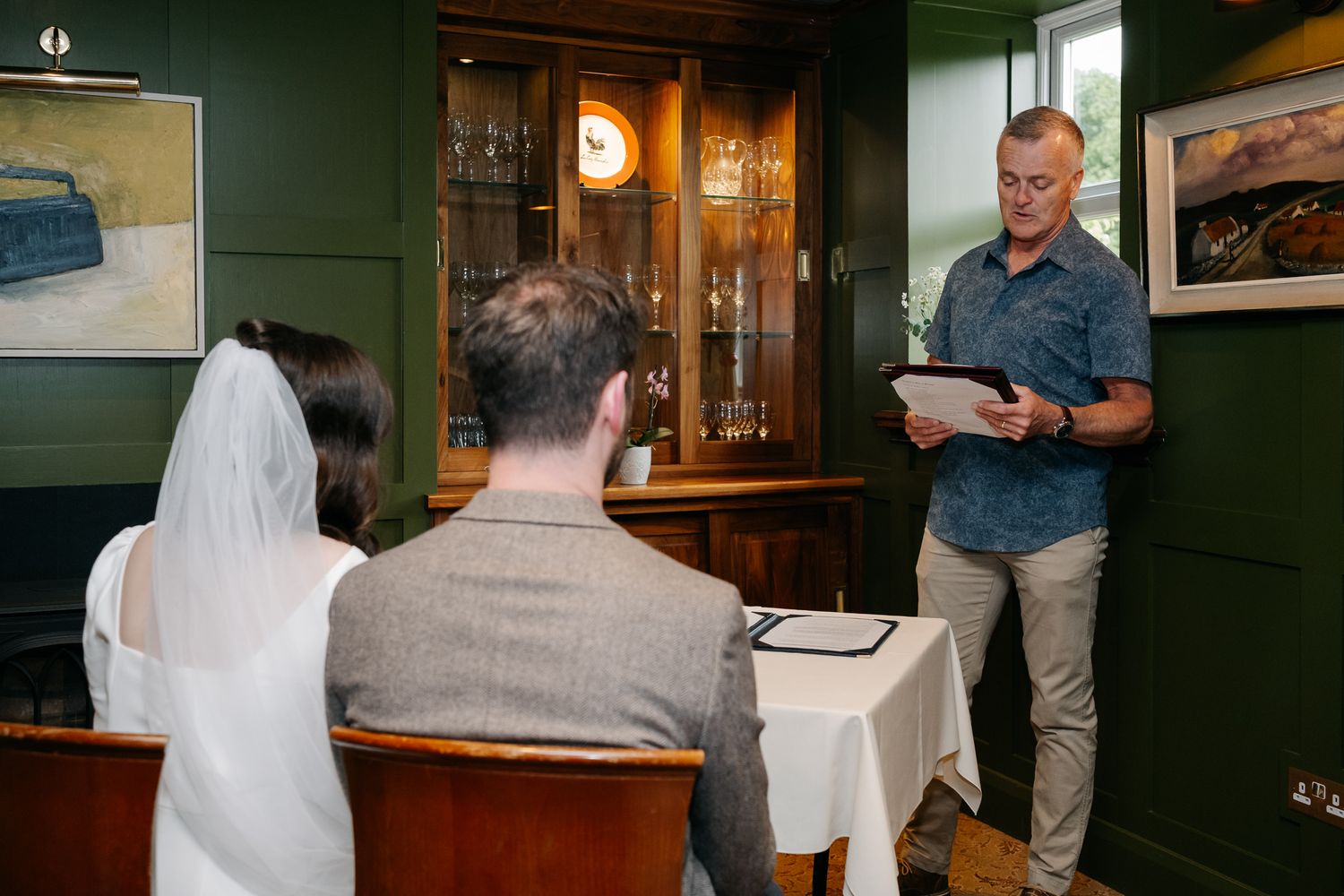 A couple sits before an officiant reading vows during an intimate wedding ceremony in a cozy green-walled venue.