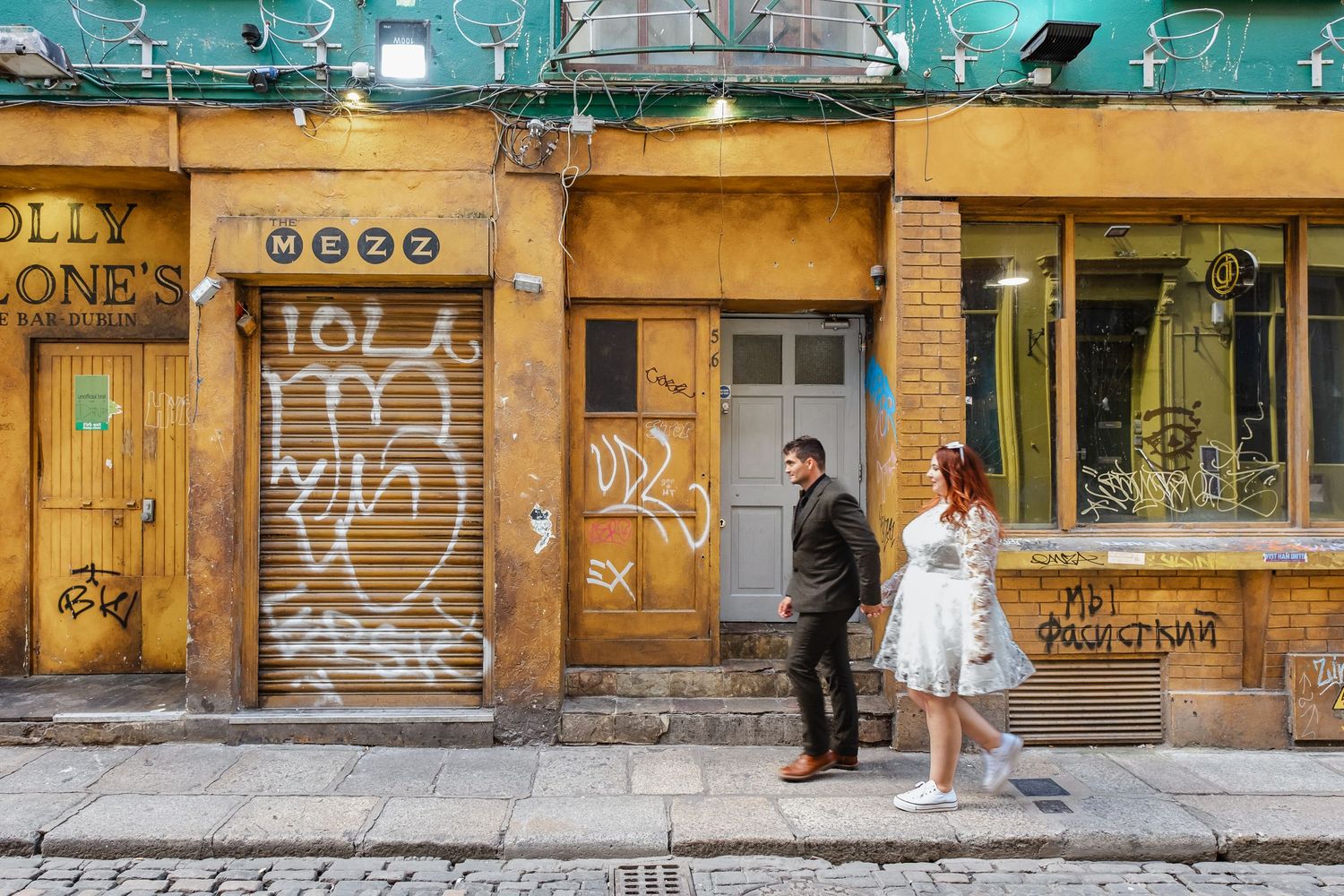 Two people standing against yellow graffitied wall in urban setting.