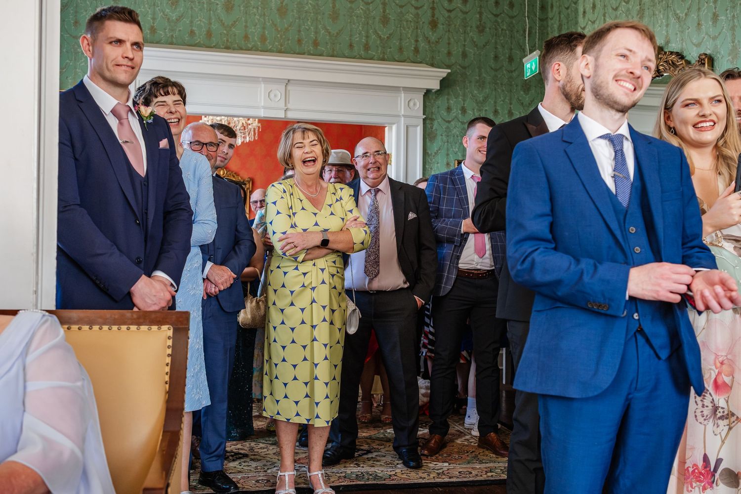 Wedding guests gathered in an elegant room with green wallpaper as someone delivers a speech.