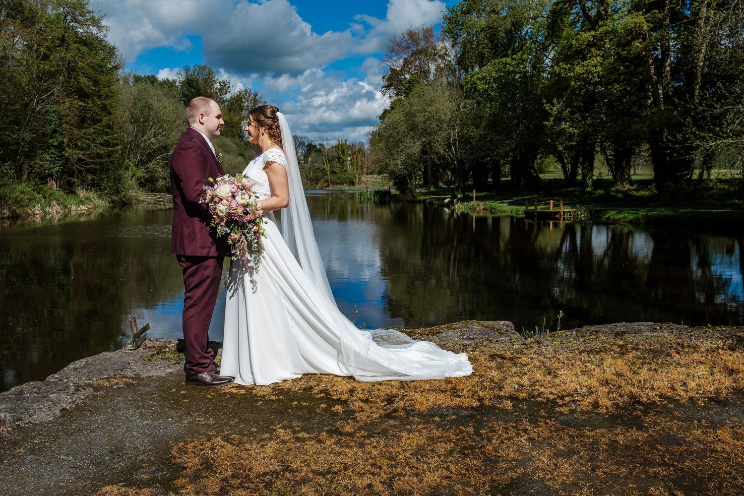 A romantic wedding couple embraces beside a serene lake surrounded by autumn trees and fallen leaves.