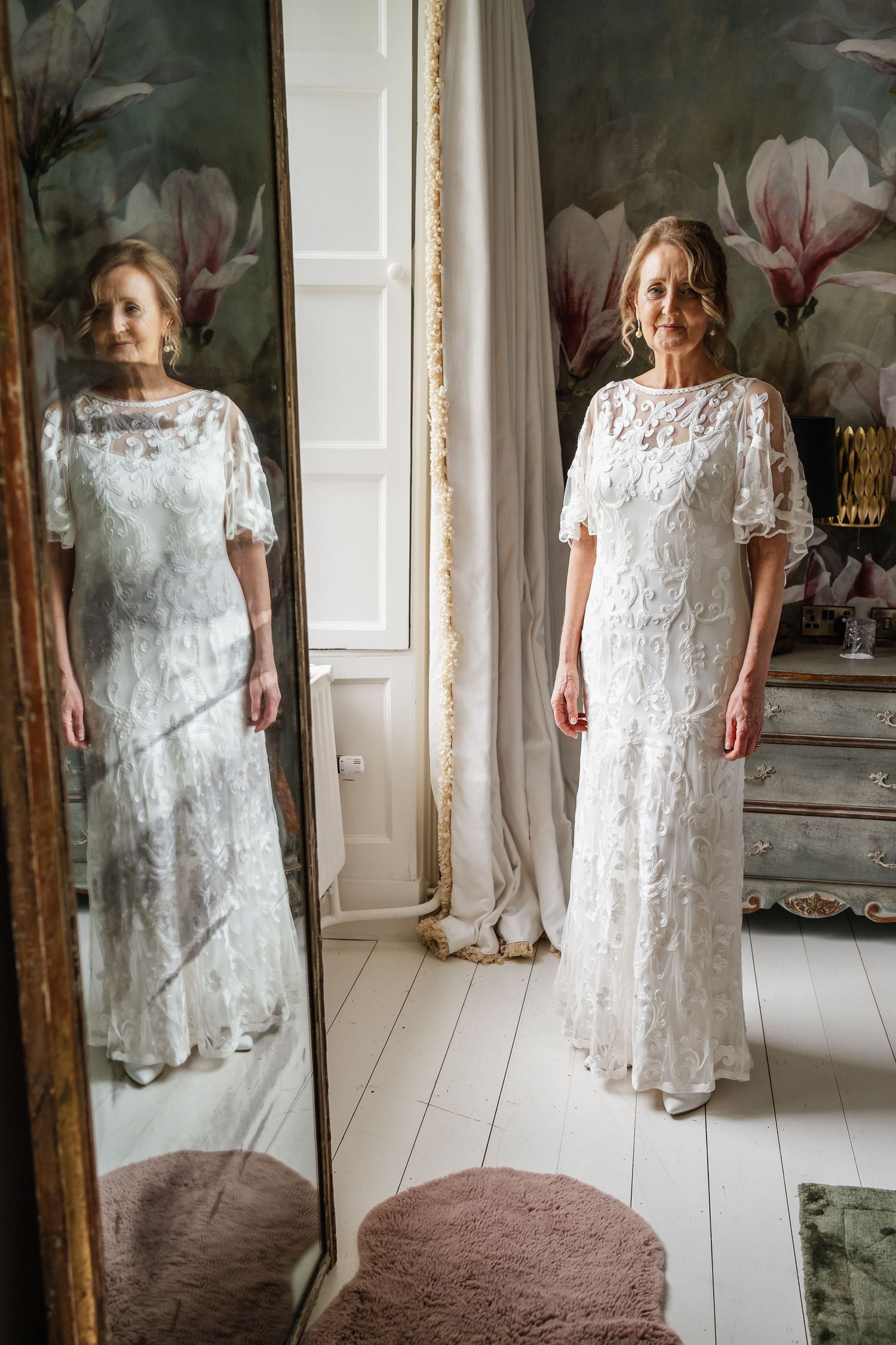 Bride in lace dress looking at reflection in mirror against floral wallpaper background.