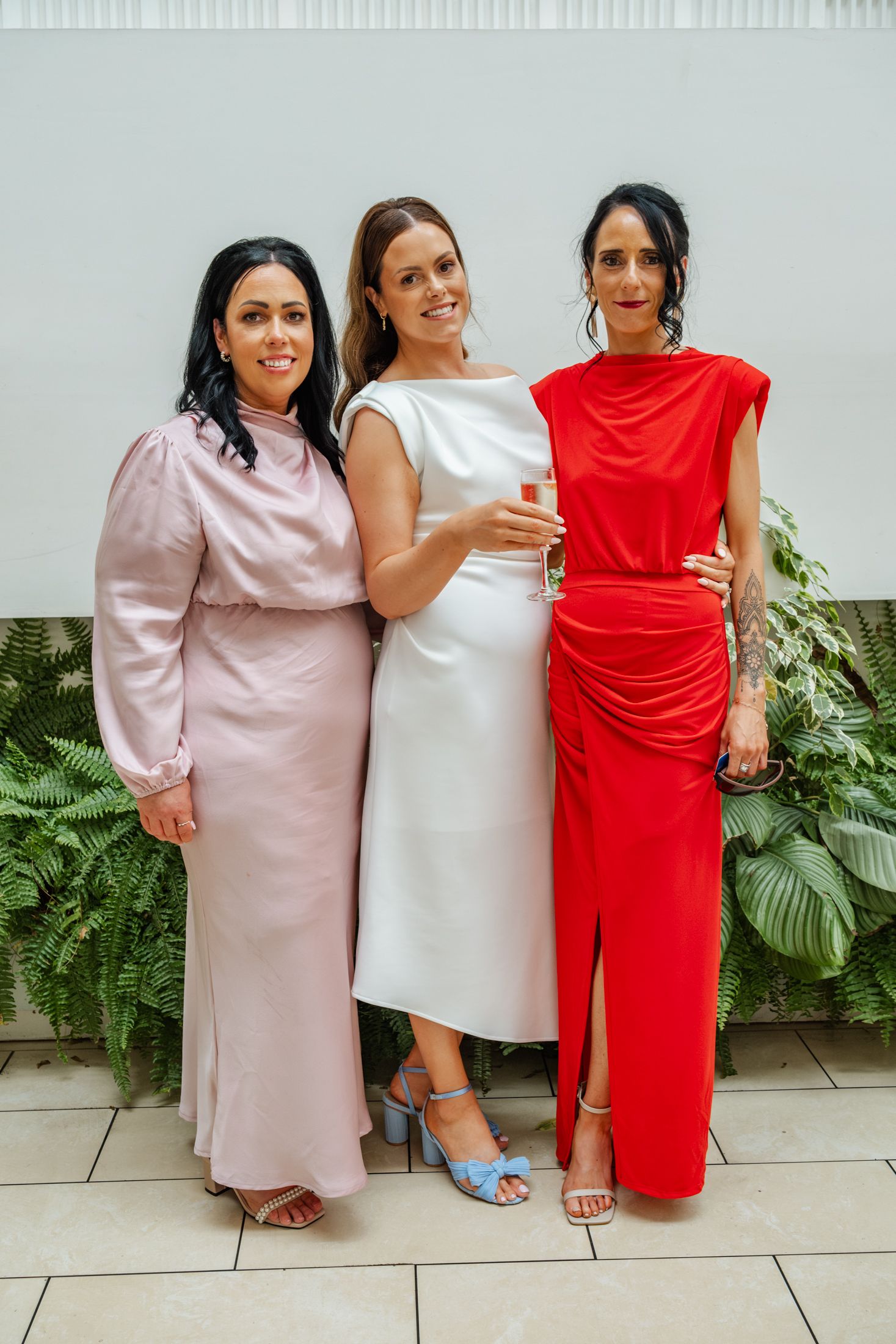 Three women in formal attire pose together against a wall with tropical plants wearing pink, white and red dresses.