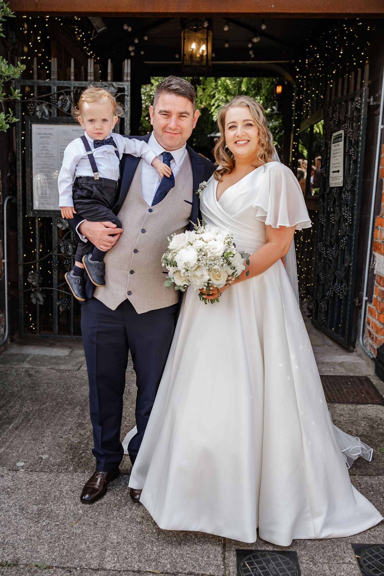 Wedding party poses under venue entrance sign holding white floral bouquet.