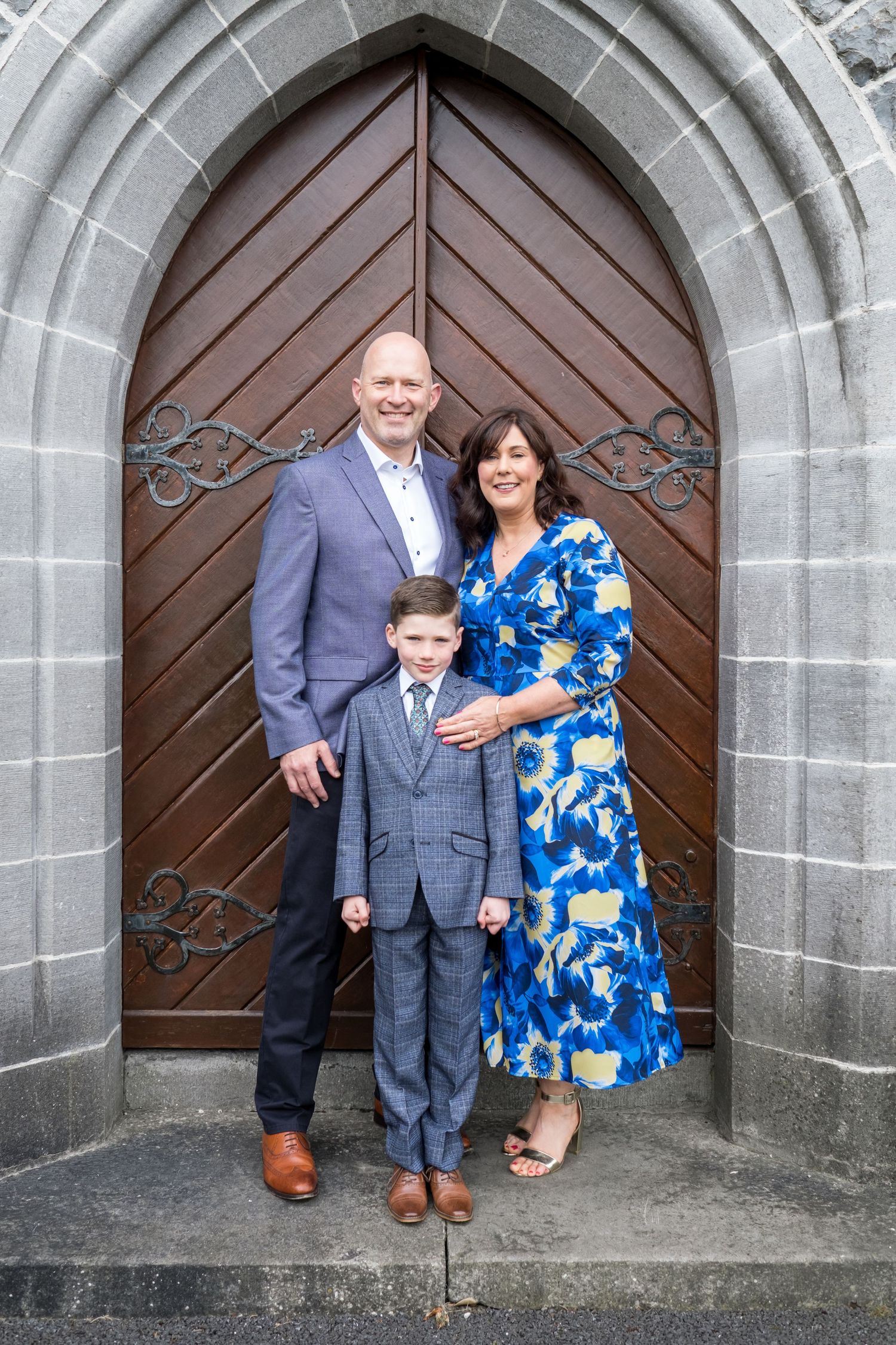 Family of three posing in front of a grand wooden arched church door, dressed in formal attire for a special occasion.