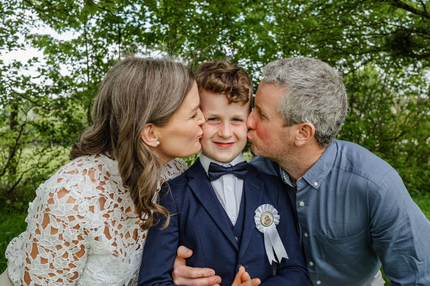 A young boy in a navy suit smiles as his parents kiss his cheeks outdoors among green trees.