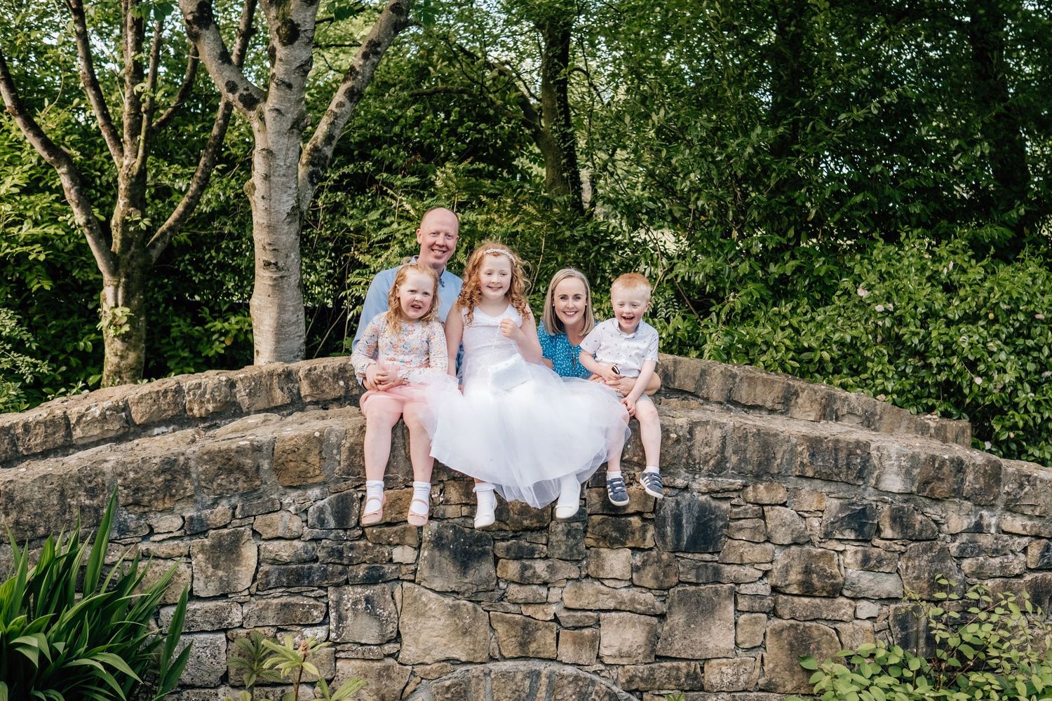 Family of five sitting on a stone bridge arch surrounded by lush green trees, smiling outdoors.