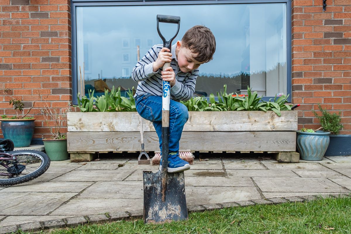 Young boy uses both feet to push a large garden spade into soil on a stone patio outdoors.