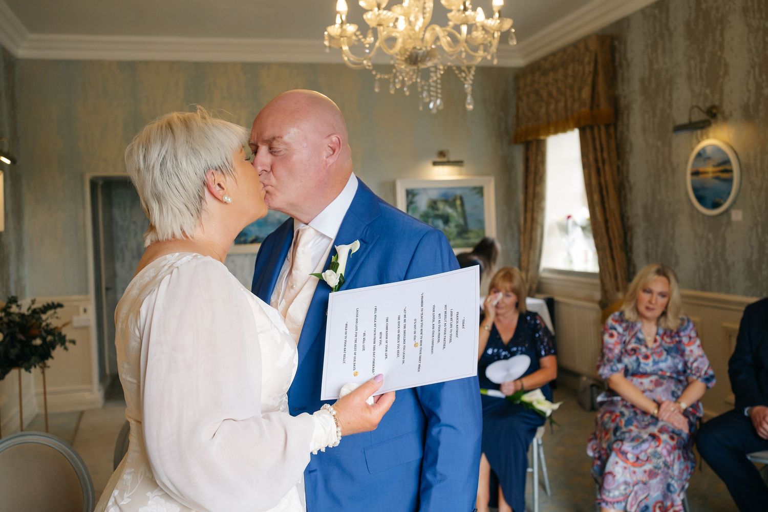 Bride and groom share first kiss during wedding ceremony, groom in blue suit holding marriage certificate.