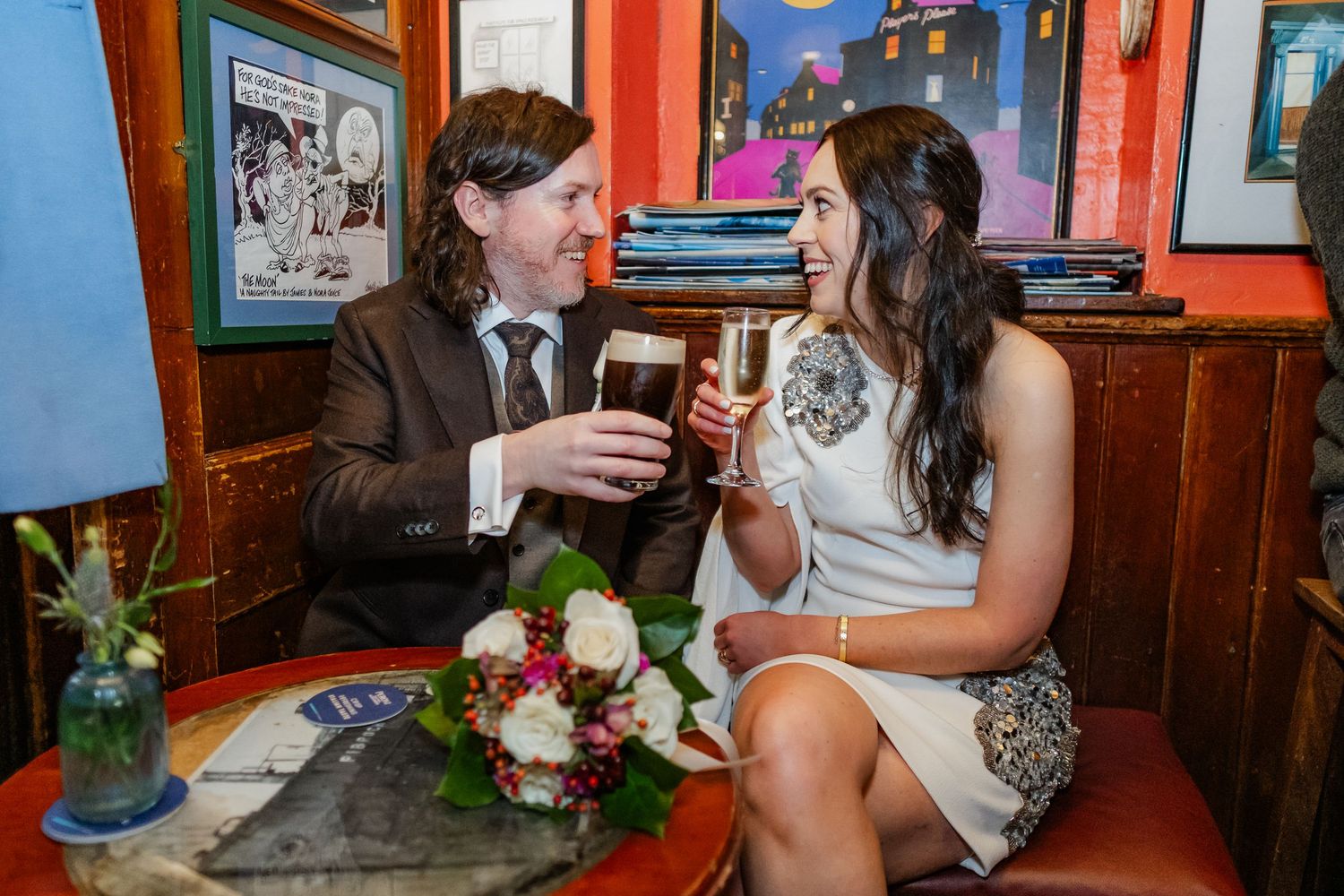 Couple in wedding attire toasting with beer glasses at a cozy pub, with a bridal bouquet on the table.