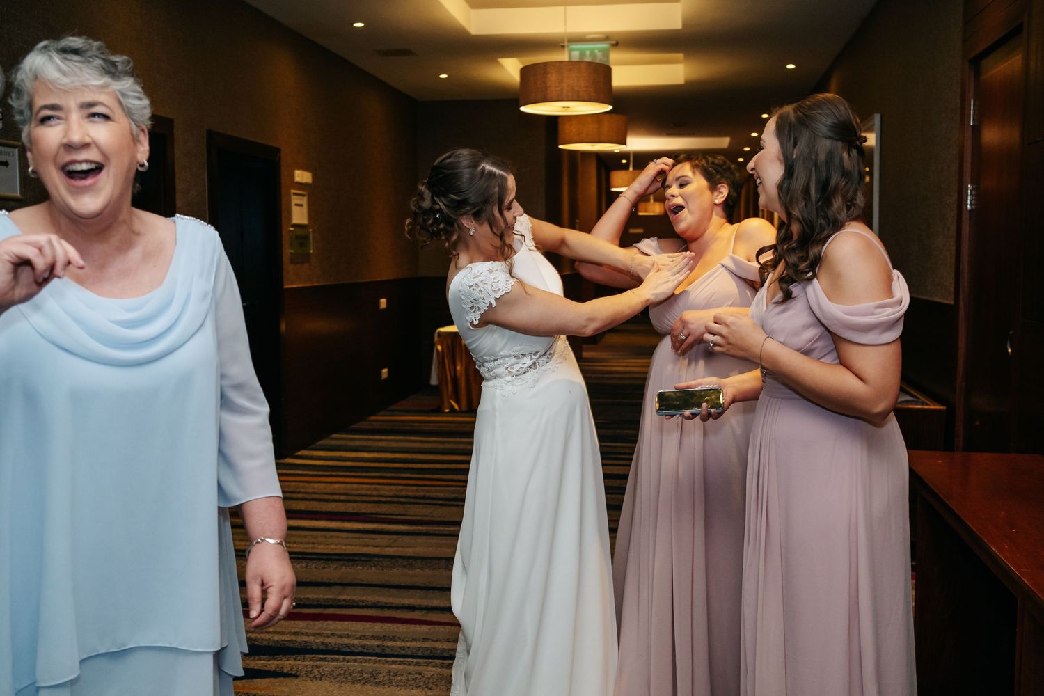 Bride laughing with bridesmaids in lavender gowns in hotel hallway as older woman reacts with joy.