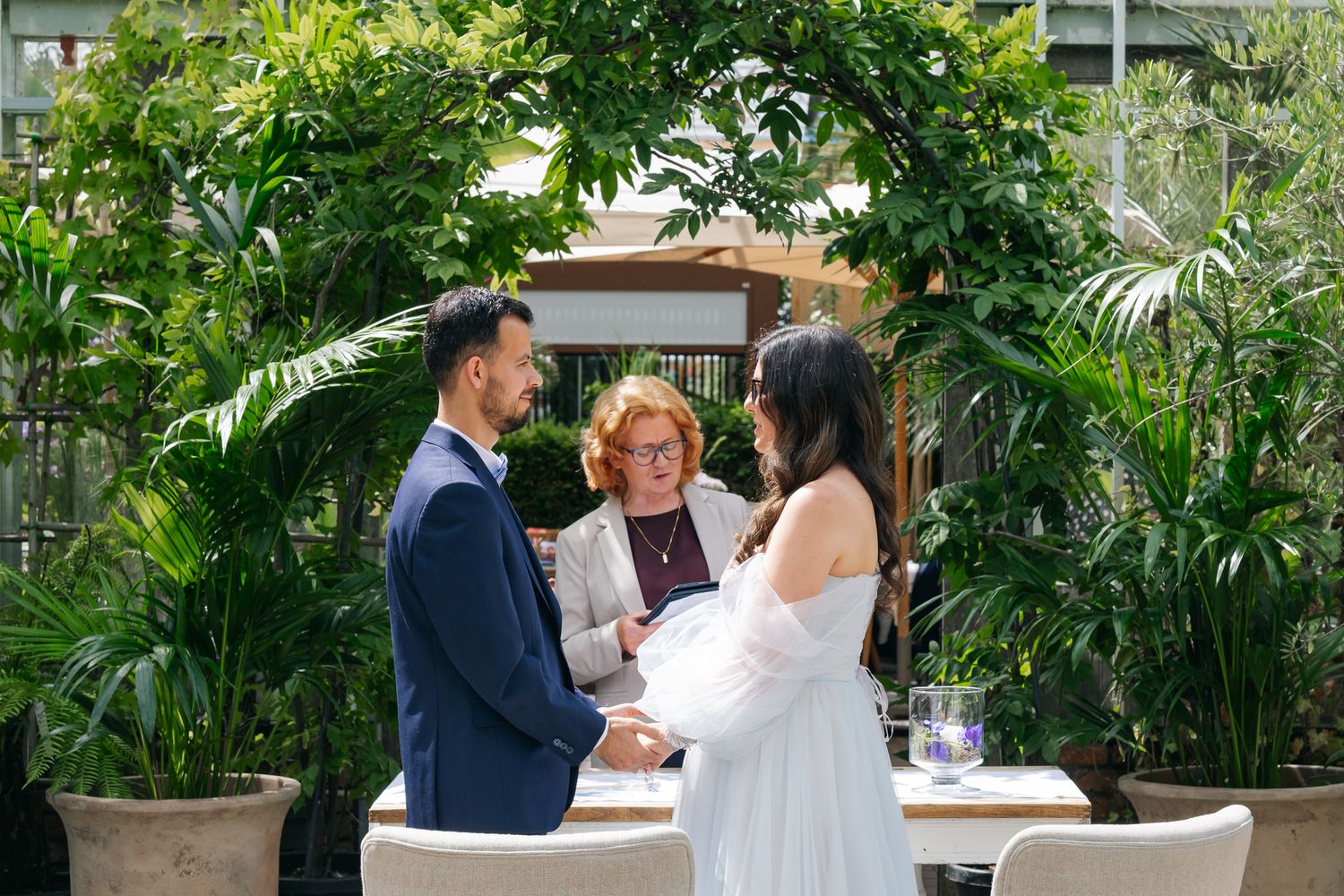 Couple exchanging vows outdoors with officiant, lush tropical greenery backdrop, elegant wedding ceremony setting.