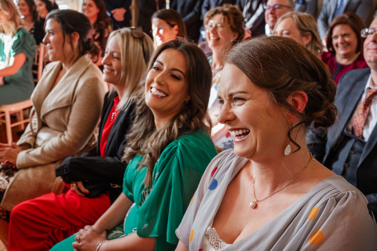 Smiling wedding guests in colorful attire laugh joyfully during an indoor ceremony.
