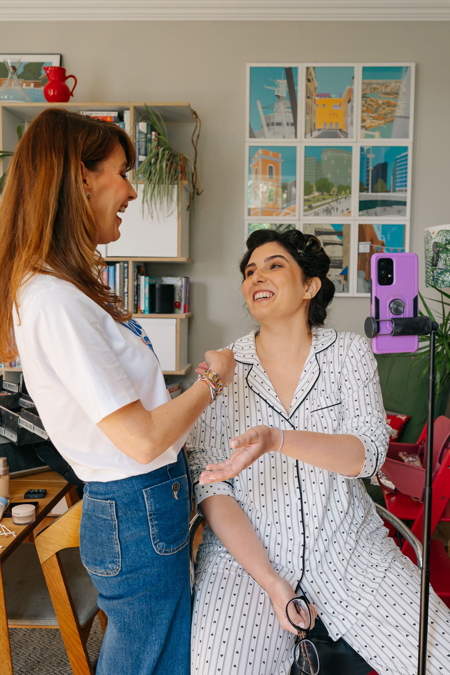 Two women smiling and chatting in a cozy room with colorful art prints on the wall behind them.