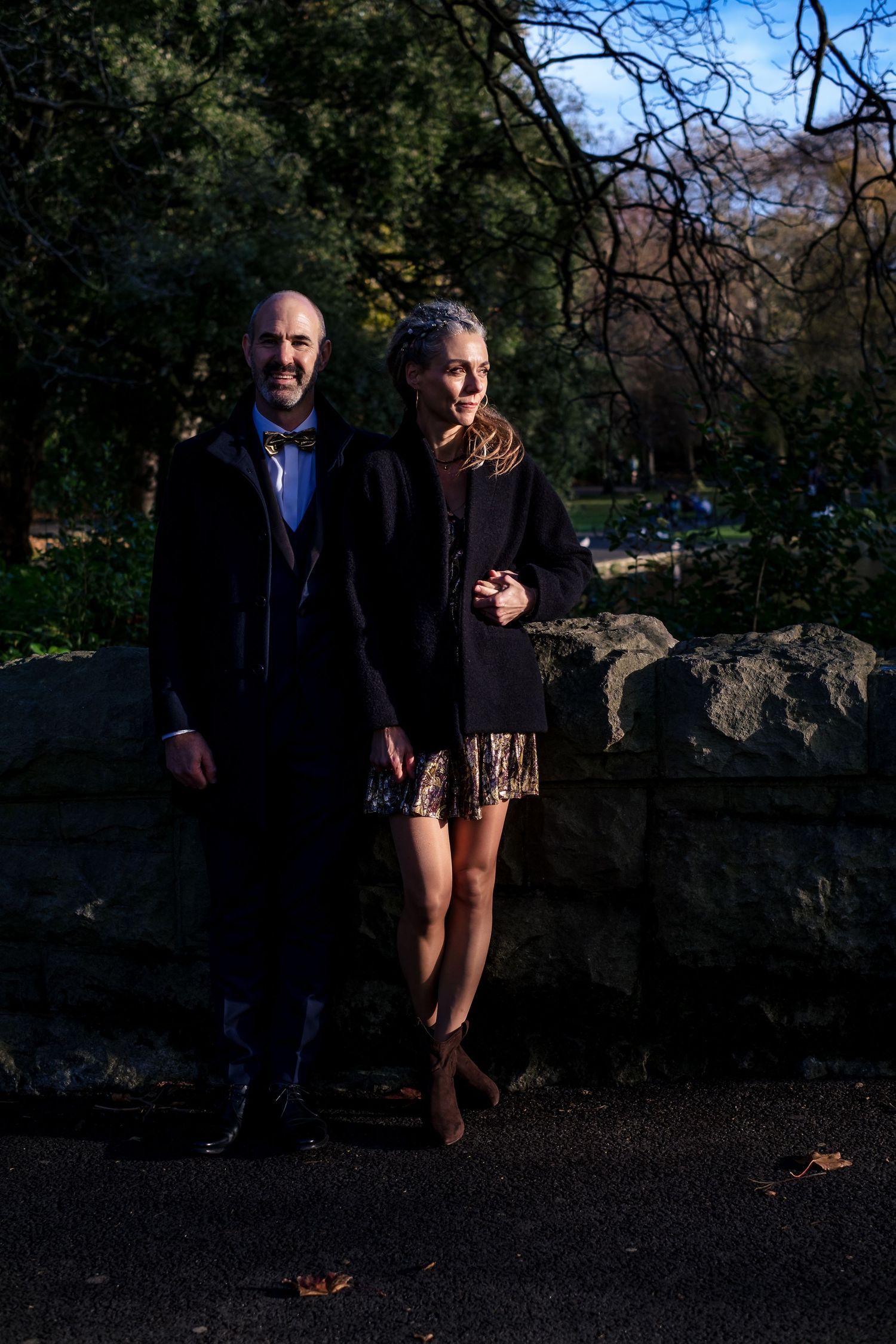 Couple posing together outdoors near a stone wall in a park, dressed in elegant dark attire on a sunny day.