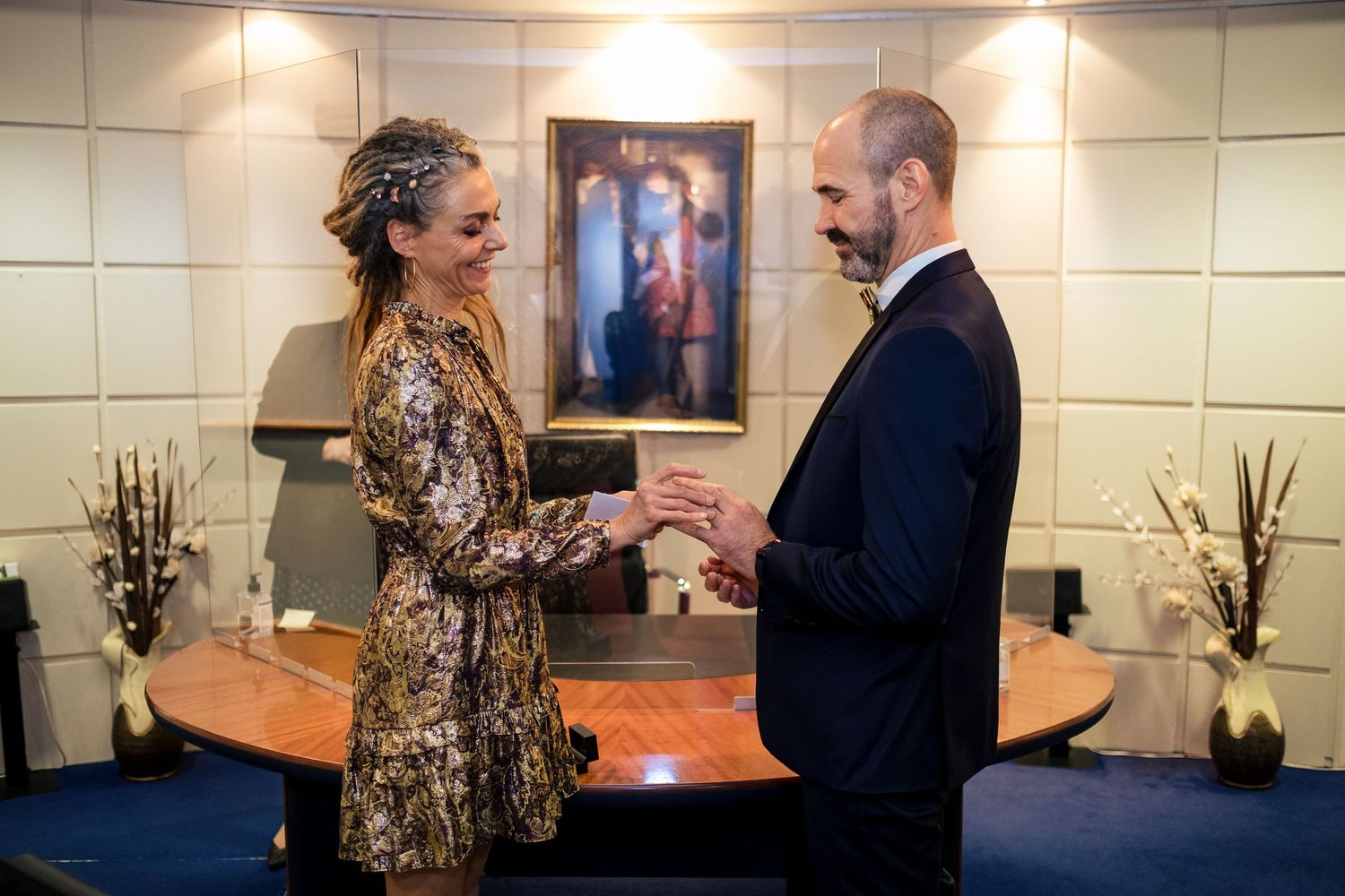 A couple exchanges rings during an intimate indoor ceremony, dressed in elegant attire with warm lighting and artwork in the background.