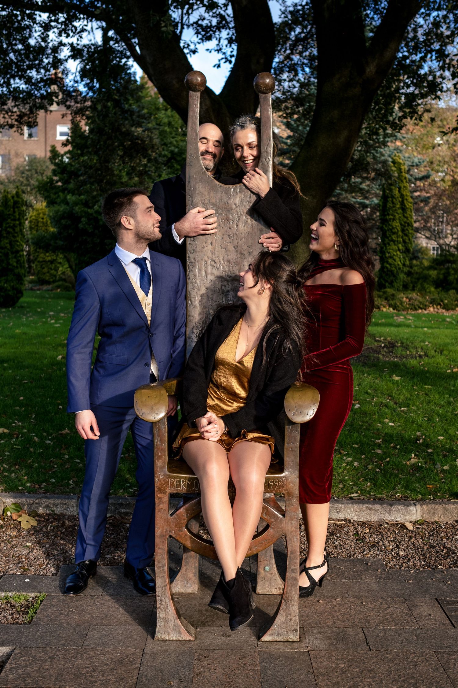 Five elegantly dressed people posing around a large wooden chair in a park setting.