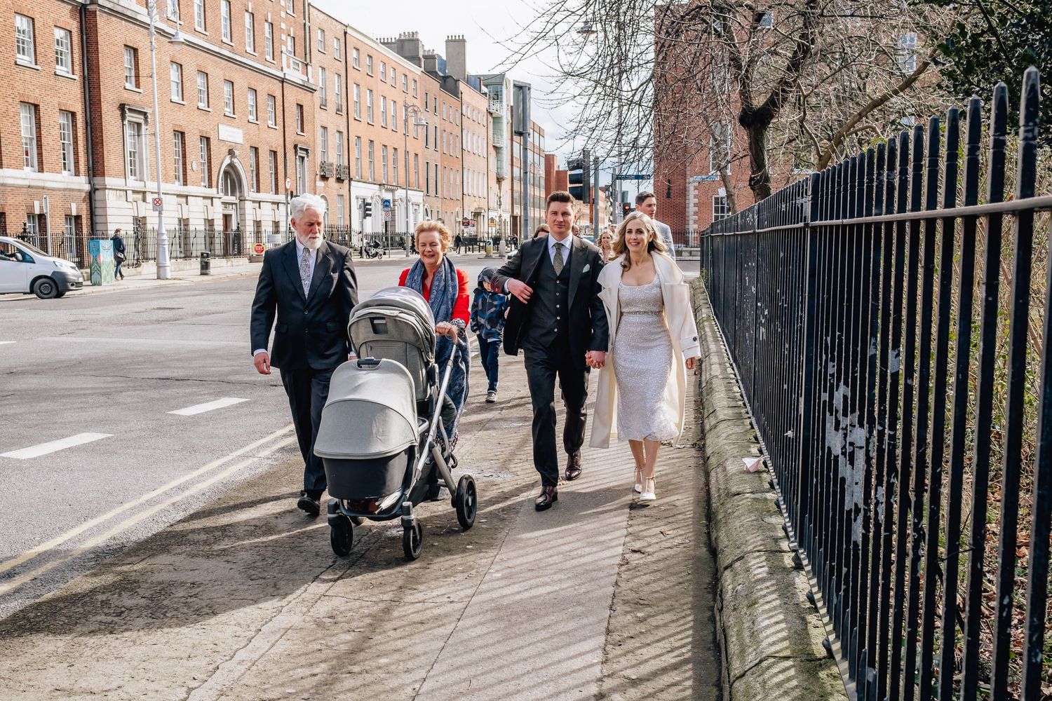 Wedding party walks down urban street with historic buildings and iron railings.