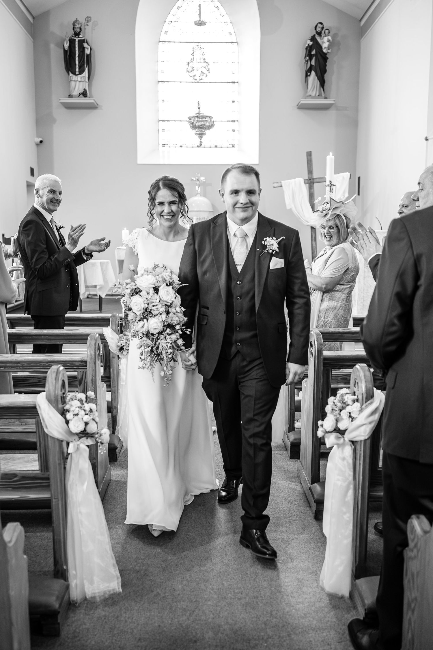 A wedding ceremony at a church interior features a couple walking down the aisle decorated with white floral arrangements.